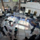 Overhead view of a spacious atrium with a central palm tree, sunlit tiled floor, and people lined up or gathered along the walls and seating areas.
