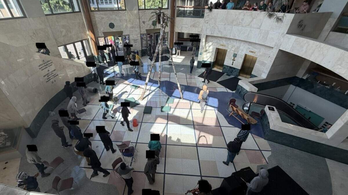 Overhead view of a spacious atrium with a central palm tree, sunlit tiled floor, and people lined up or gathered along the walls and seating areas.