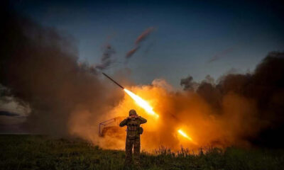 Soldier in camouflage in a field as a rocket launcher fires, leaving a bright orange flame and dense smoke behind him at dusk/ dawn.