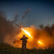 Soldier in camouflage in a field as a rocket launcher fires, leaving a bright orange flame and dense smoke behind him at dusk/ dawn.