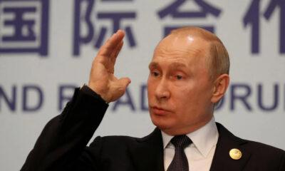 Man in a dark suit and tie speaks at a conference, raising his right hand near his temple against a backdrop of large lettering behind him.