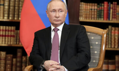 Man in a dark suit and pink tie seated in a wooden chair in a library, with a red and blue flag behind him.