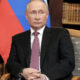 Man in a dark suit and pink tie seated in a wooden chair in a library, with a red and blue flag behind him.