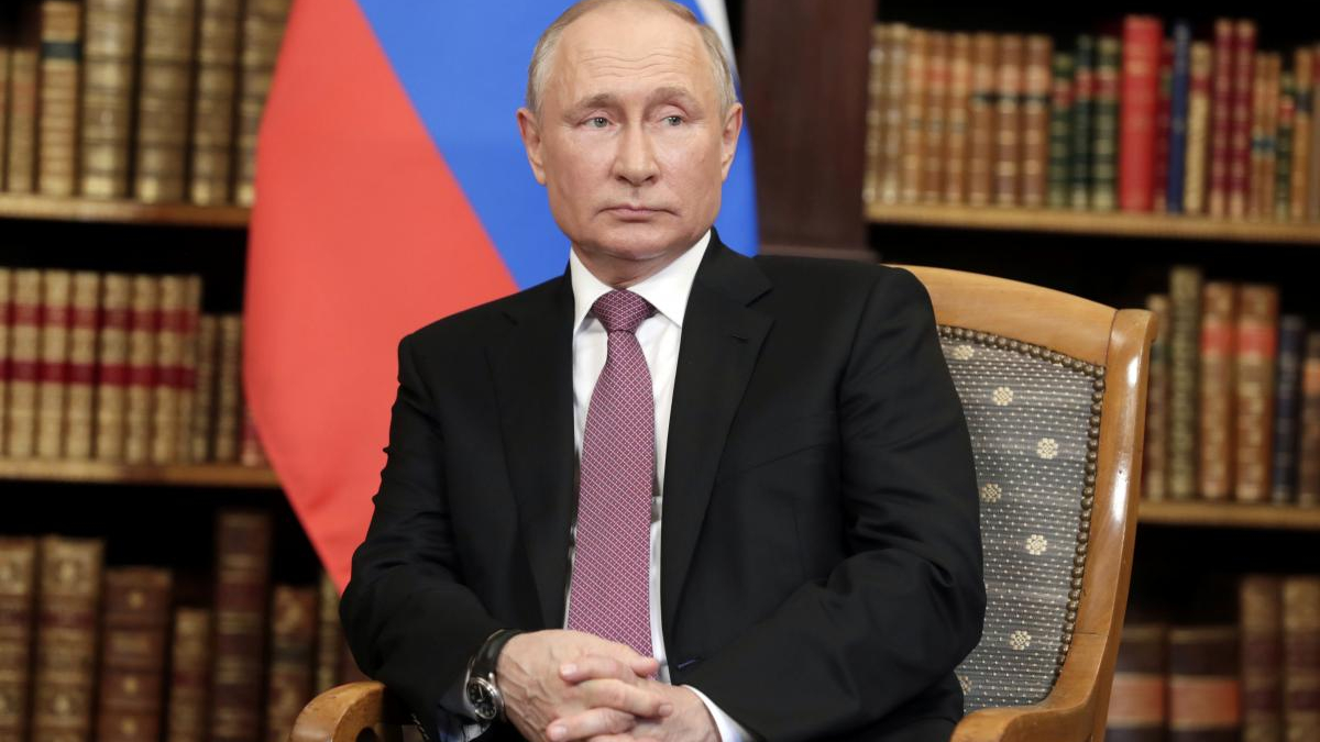 Man in a dark suit and pink tie seated in a wooden chair in a library, with a red and blue flag behind him.