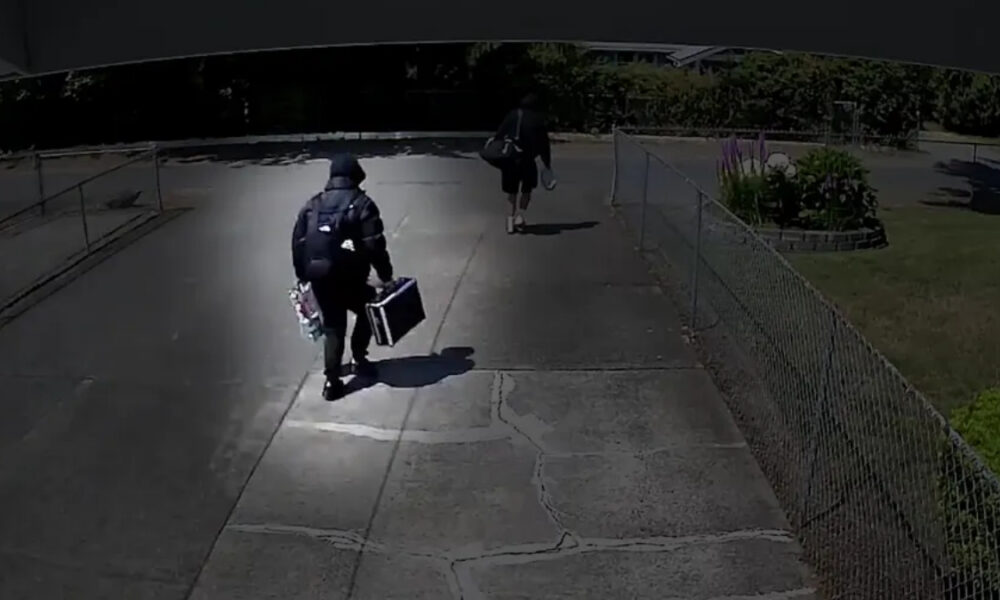 Two people walk away from the camera along a concrete path; one wears a backpack and carries a rectangular case, the other carries a bag and holds items in one hand.