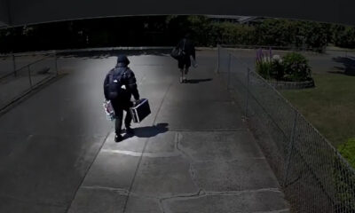 Two people walk away from the camera along a concrete path; one wears a backpack and carries a rectangular case, the other carries a bag and holds items in one hand.