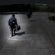 Two people walk away from the camera along a concrete path; one wears a backpack and carries a rectangular case, the other carries a bag and holds items in one hand.