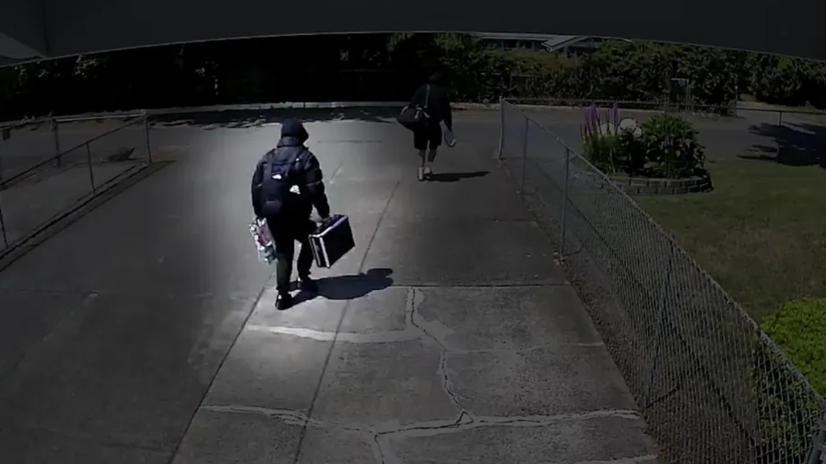 Two people walk away from the camera along a concrete path; one wears a backpack and carries a rectangular case, the other carries a bag and holds items in one hand.
