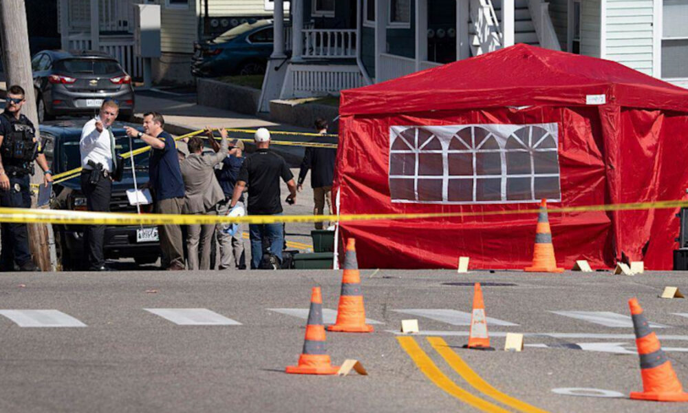 Police and investigators stand behind yellow caution tape on a residential street, near a red tent and traffic cones marking a crime scene.