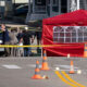 Police and investigators stand behind yellow caution tape on a residential street, near a red tent and traffic cones marking a crime scene.