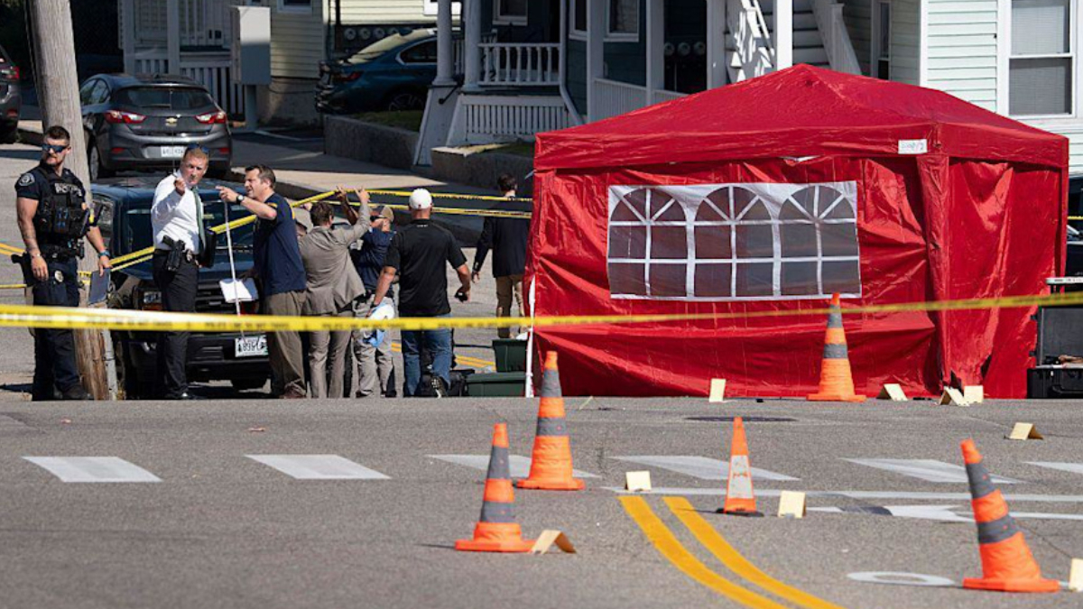 Police and investigators stand behind yellow caution tape on a residential street, near a red tent and traffic cones marking a crime scene.