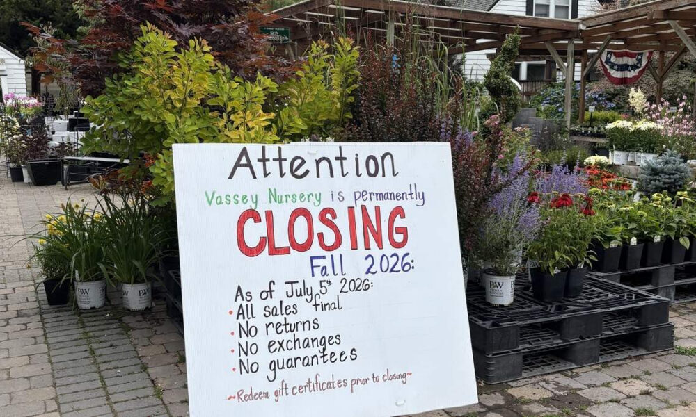 Sign at a plant nursery announcing permanent closing in Fall 2026, with bullet points about sales, returns, exchanges, and guarantees, surrounded by potted plants and flowers.