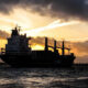 Silhouette of a large cargo ship with cranes on choppy water at sunset, orange sky behind clouds.