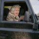 Older woman wearing a patterned headscarf driving a black Land Rover, mud splattered on the door.