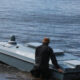 Person wearing a brown hat helps push a gray speedboat into calm water at the lake, shoreline in view.