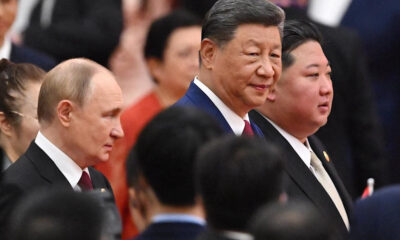 Three men in dark suits at a formal event, the central figure with a slight smile, others nearby in profile.