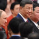 Three men in dark suits at a formal event, the central figure with a slight smile, others nearby in profile.