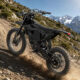 Black electric dirt bike on a rocky mountain trail with snow-capped peaks in the distance.