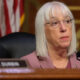 Senator Durbin seated at a hearing table with a microphone, wearing a pink blazer and glasses, nameplate visible in the foreground.