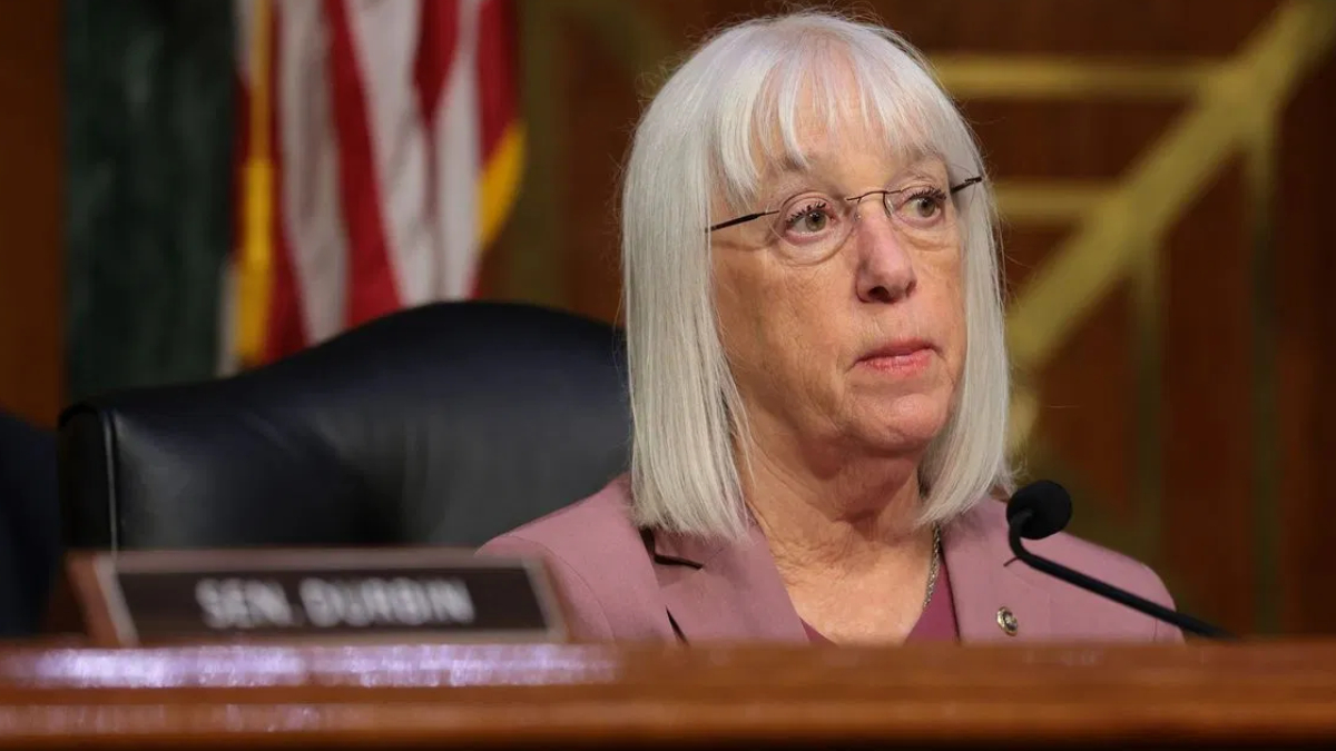 Senator Durbin seated at a hearing table with a microphone, wearing a pink blazer and glasses, nameplate visible in the foreground.