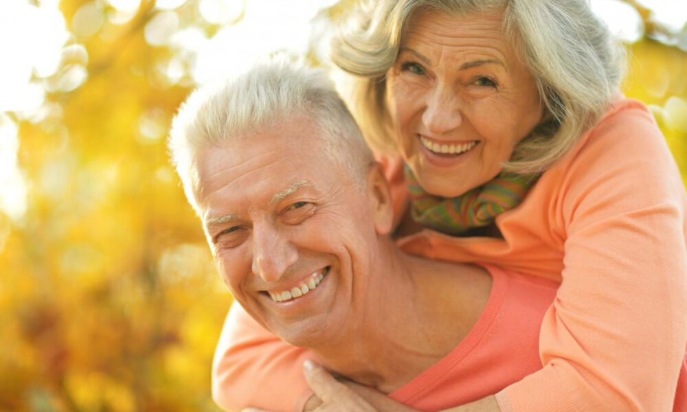 Smiling elderly man carrying a joyful woman on his back outdoors in warm autumn light.