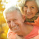 Smiling elderly man carrying a joyful woman on his back outdoors in warm autumn light.