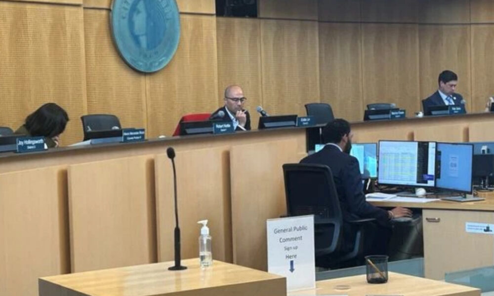 Council chamber with three officials at a curved desk; a man in the center speaks into a microphone, monitors on the desk nearby.