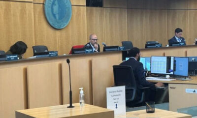 Council chamber with three officials at a curved desk; a man in the center speaks into a microphone, monitors on the desk nearby.