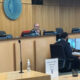 Council chamber with three officials at a curved desk; a man in the center speaks into a microphone, monitors on the desk nearby.