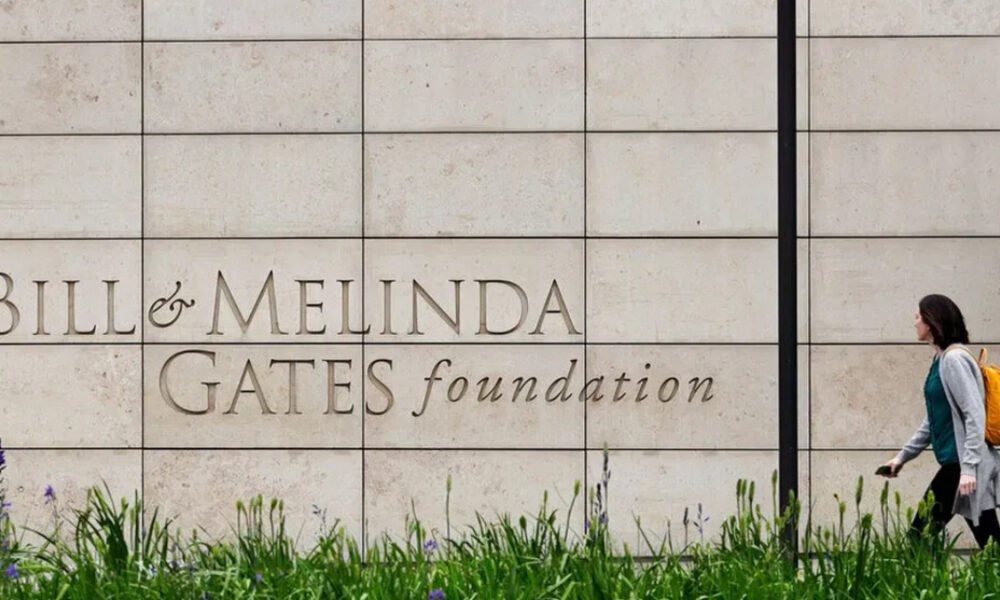 A person with an orange backpack walks past a stone wall engraved with 'Bill & Melinda Gates Foundation' text on the wall.