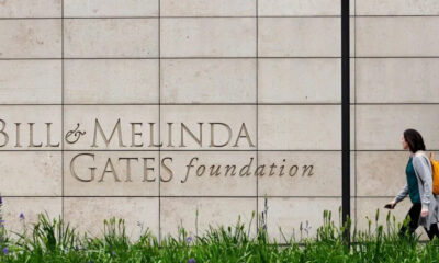 A person with an orange backpack walks past a stone wall engraved with 'Bill & Melinda Gates Foundation' text on the wall.