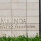 A person with an orange backpack walks past a stone wall engraved with 'Bill & Melinda Gates Foundation' text on the wall.