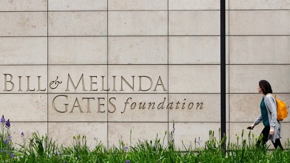 A person with an orange backpack walks past a stone wall engraved with 'Bill & Melinda Gates Foundation' text on the wall.