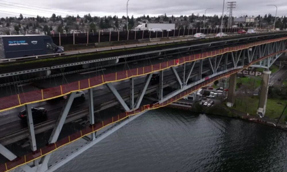 Two-level steel bridge over a river with moving traffic on the upper deck and a red pedestrian walkway with yellow railing along the side.