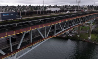 Two-level steel bridge over a river with moving traffic on the upper deck and a red pedestrian walkway with yellow railing along the side.