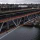 Two-level steel bridge over a river with moving traffic on the upper deck and a red pedestrian walkway with yellow railing along the side.