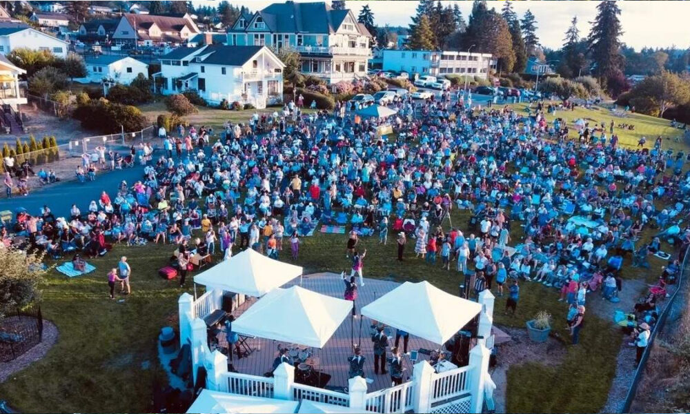 Outdoor community event with a crowd seated on a lawn around a small stage under white tents, in a residential neighborhood.