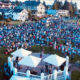 Outdoor community event with a crowd seated on a lawn around a small stage under white tents, in a residential neighborhood.