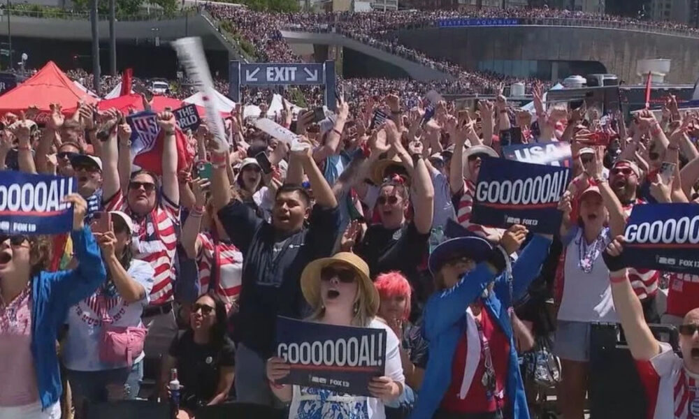 Large stadium crowd cheering and waving signs that read GOOOOOOAL, celebrating a goal with people of various ages and outfits