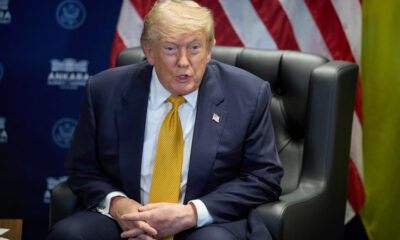 Man in a dark suit and yellow tie speaks at a meeting, with an American flag behind him.