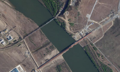 Aerial view of a river or canal with a long red bridge crossing it, surrounded by fields and a few buildings on either side.