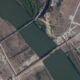 Aerial view of a river or canal with a long red bridge crossing it, surrounded by fields and a few buildings on either side.