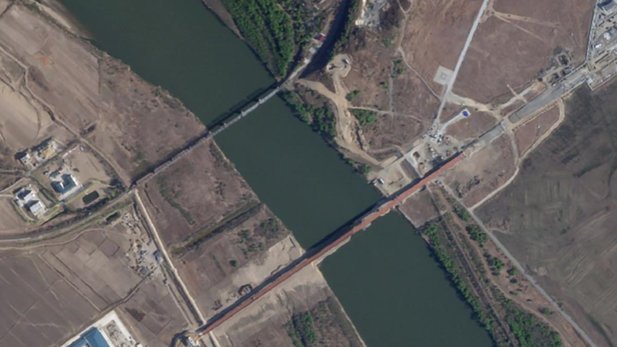 Aerial view of a river or canal with a long red bridge crossing it, surrounded by fields and a few buildings on either side.