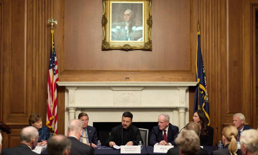 Formal panel discussion in a wood-paneled room, with an American flag and another flag flanking a marble fireplace and a framed portrait above.