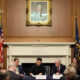 Formal panel discussion in a wood-paneled room, with an American flag and another flag flanking a marble fireplace and a framed portrait above.