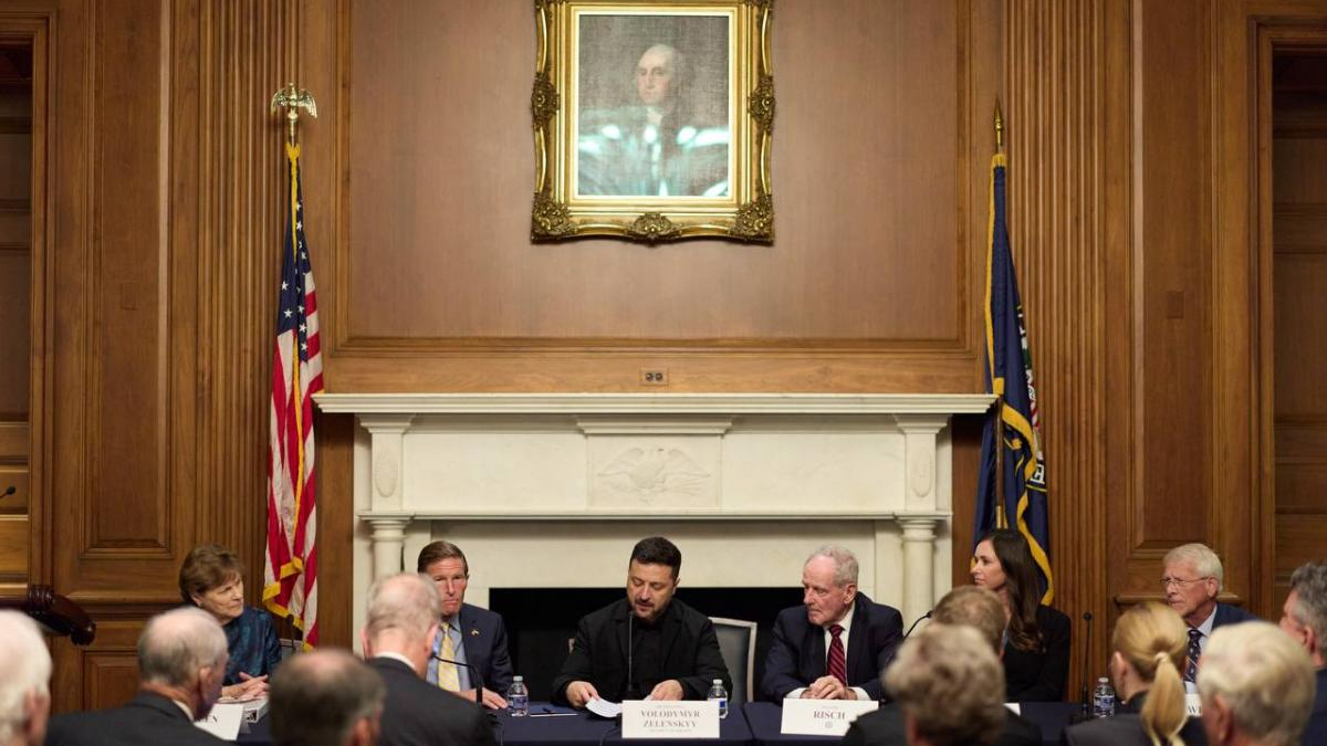 Formal panel discussion in a wood-paneled room, with an American flag and another flag flanking a marble fireplace and a framed portrait above.