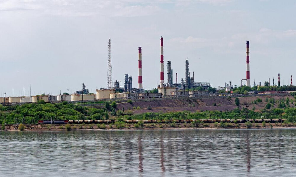 Industrial refinery with tall red-and-white smokestacks along a riverbank, under a clear sky