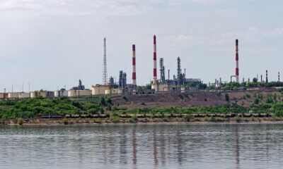 Industrial refinery with tall red-and-white smokestacks along a riverbank, under a clear sky