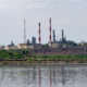 Industrial refinery with tall red-and-white smokestacks along a riverbank, under a clear sky