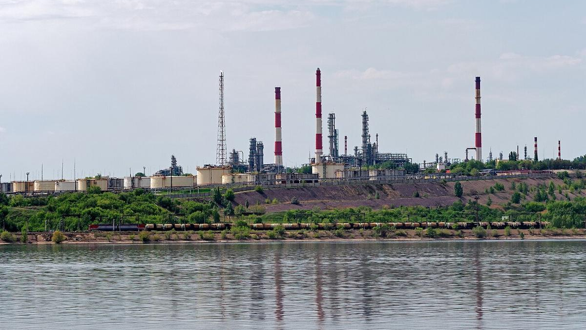 Industrial refinery with tall red-and-white smokestacks along a riverbank, under a clear sky